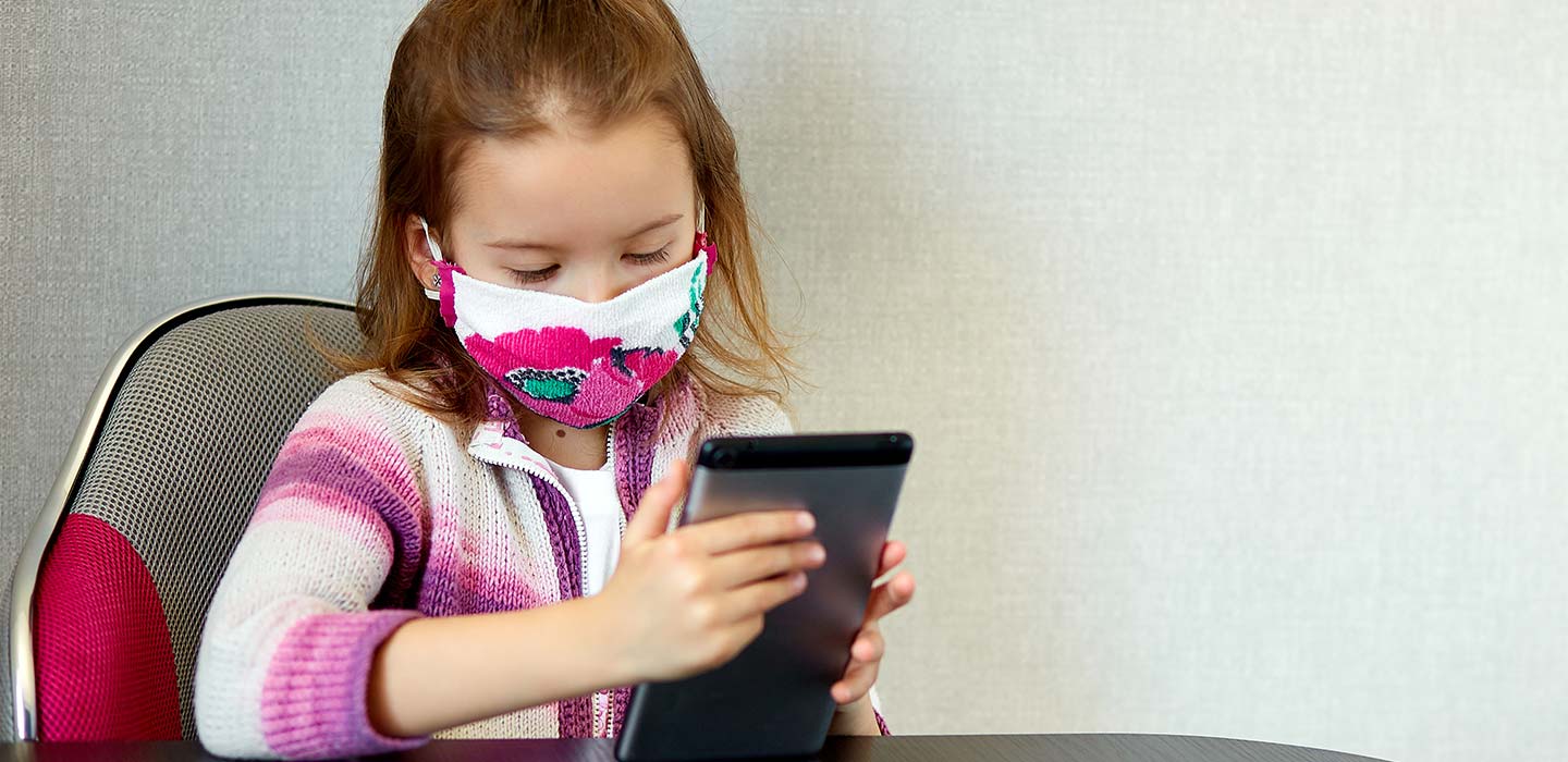 Niña niña en casa, usando una máscara de protección y juegos en una tableta, cuarentena Coronavirus.